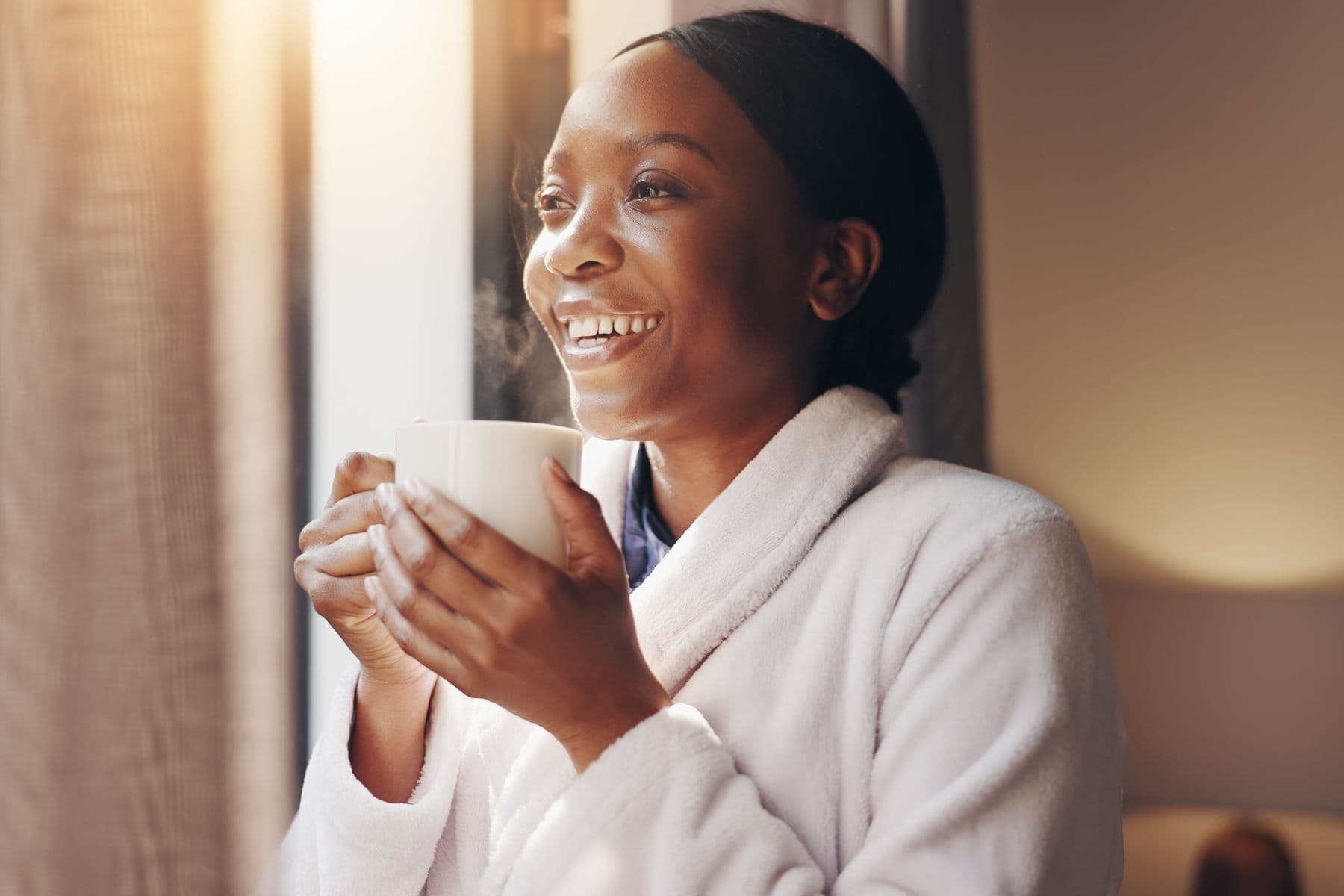 A woman in a robe holding a warm drink by a window in morning light