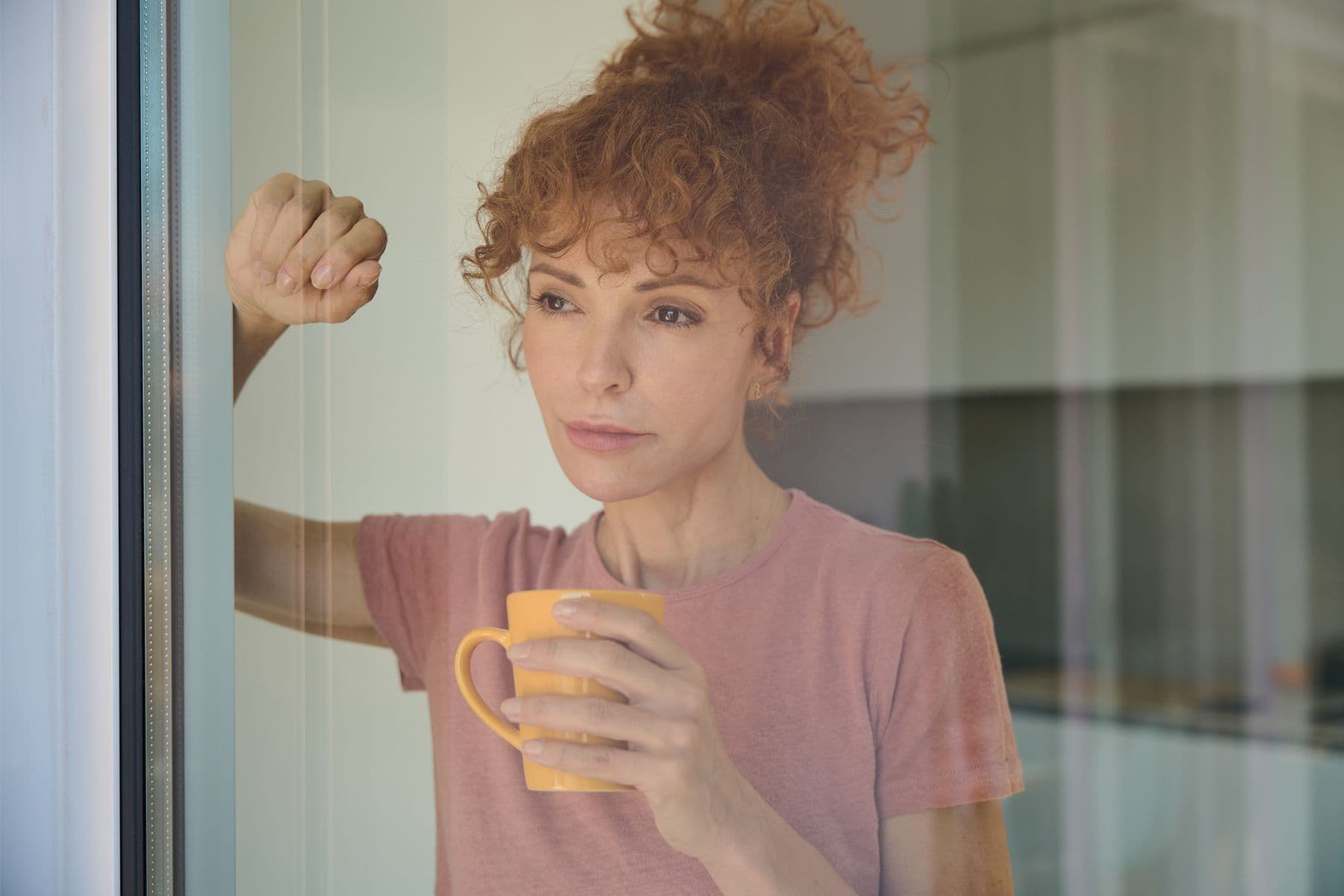 A woman standing at a window holding a mug, looking out